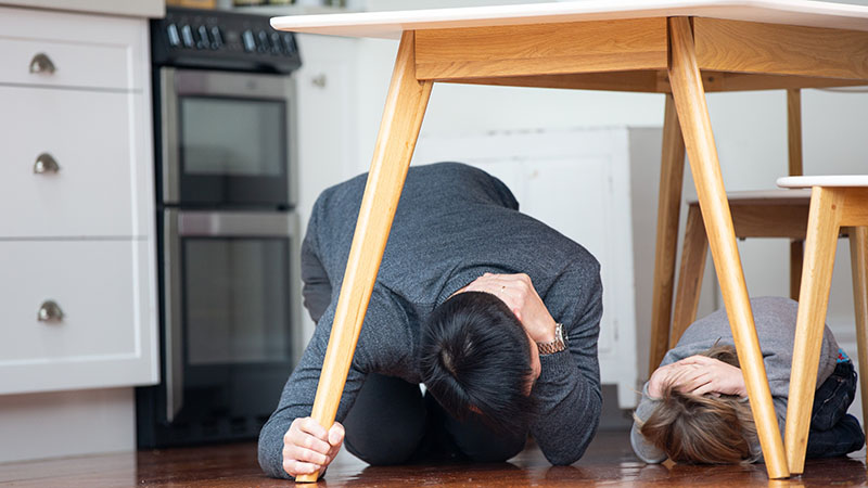 A dad and child doing Drop Cover and Hold under a dining table