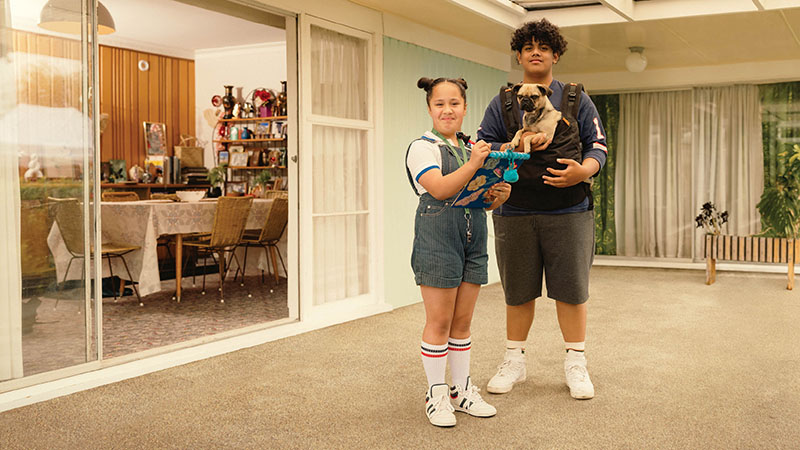 A young girl with a clipboard and her brother with a dog in a front pack outside their house, staring at the viewer